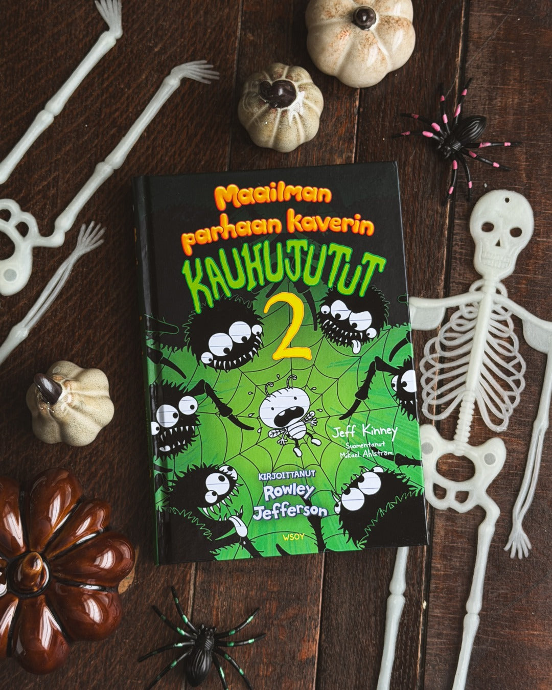 Jeff Kinneyn Maailman parhaan kaverin kauhujutut 2 -kirja makaa tummalla puupöydällä. Kirjan ympärillä on muovisia luurankoja ja hämähäkkejä sekä pieniä koristekurpitsoja.
