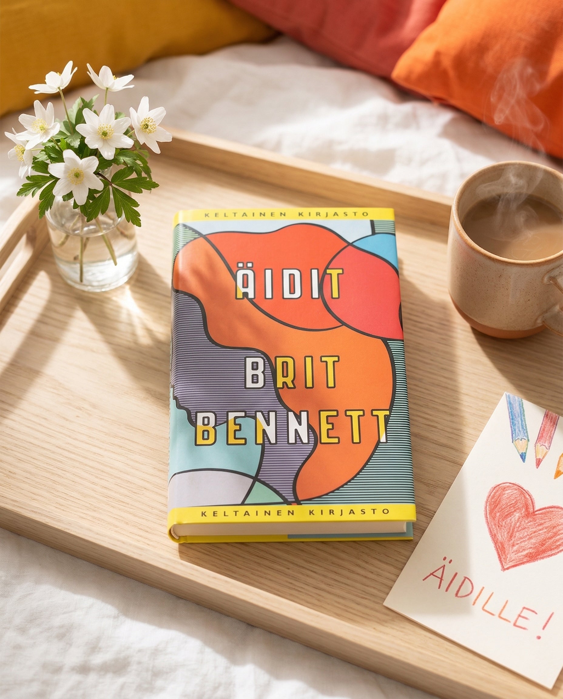 Tarjottimella on kirja Äidit, tekijä Brit Bennett. Sen vieressä on pieni maljakko valkoisilla kukilla, höyryävä kahvikuppi ja kortti, jossa on piirretty sydän. Taustalla näkyy tyynyjä.