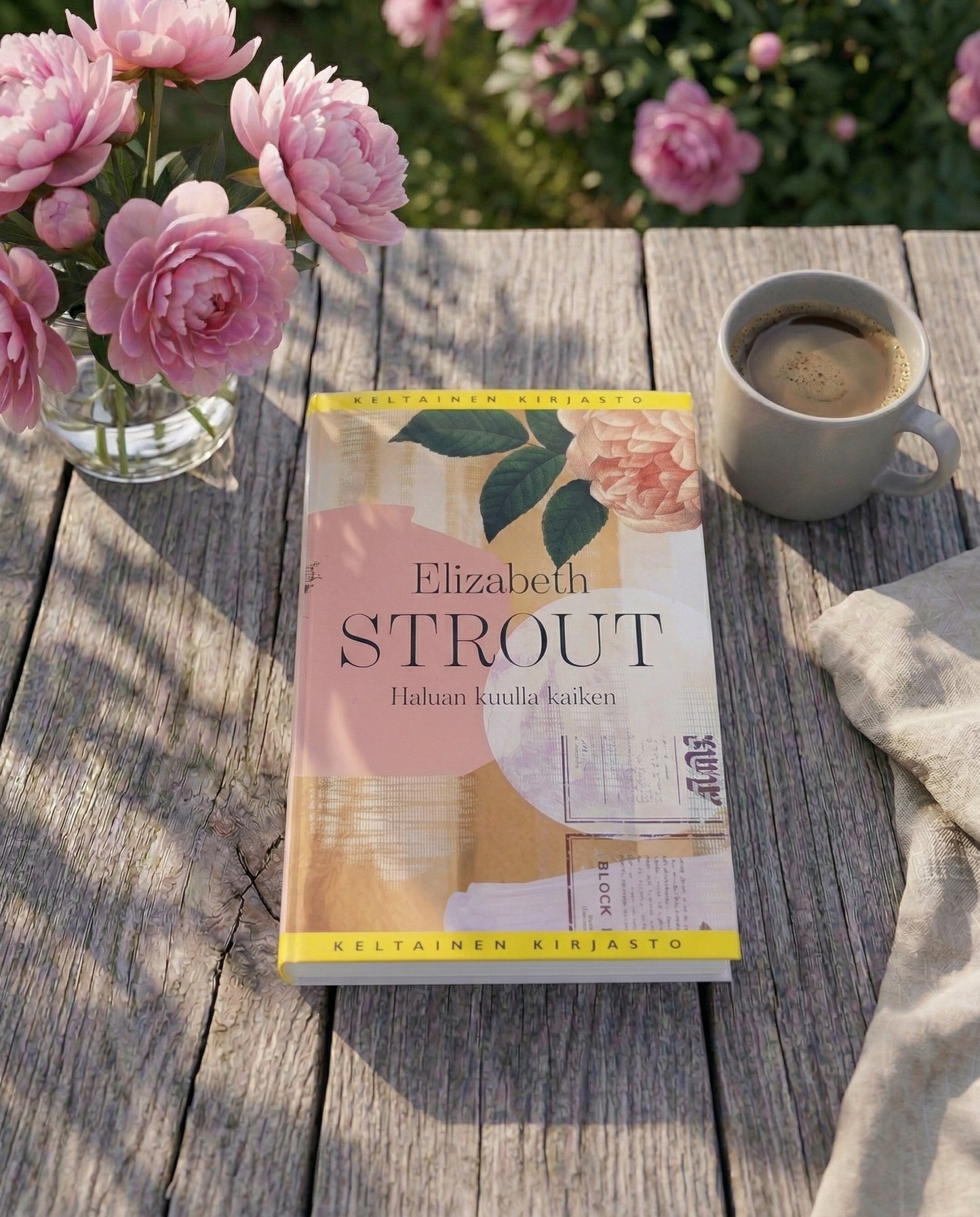 Elizabeth Stroutin kirja Haluan kuulla kaiken on asetettu puutarhapöydälle. Kirjan vieressä on kahvikuppi, kangasliina ja maljakossa vaaleanpunaisia kukkia. Taustalla näkyy kukkiva puutarha.