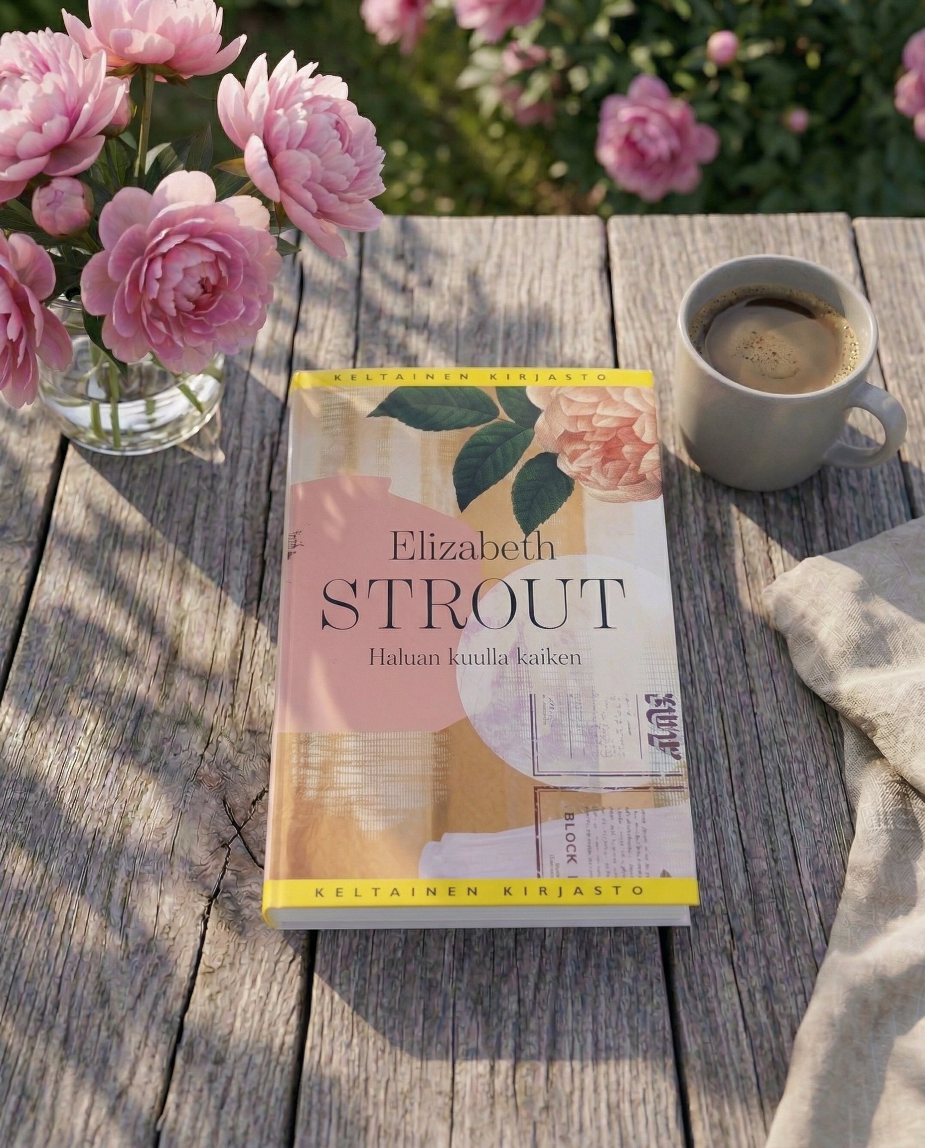 Elizabeth Stroutin kirja Haluan kuulla kaiken on asetettu puutarhapöydälle. Kirjan vieressä on kahvikuppi, kangasliina ja maljakossa vaaleanpunaisia kukkia. Taustalla näkyy kukkiva puutarha.