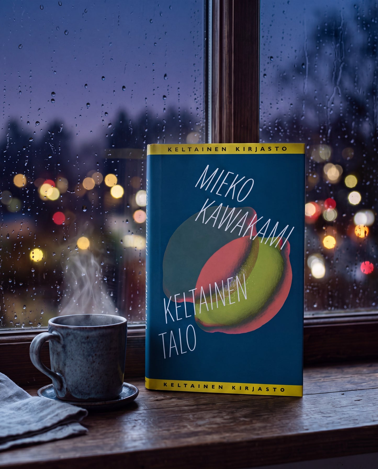 Mieko Kawakamin Keltainen talo -kirja seisoo puisella ikkunalaudalla höyryävän kupin vieressä. Kannen tummansinisellä pohjalla on vihreä hedelmä ja tekstit ”Mieko Kawakami”, ”Keltainen talo” sekä ”Keltainen kirjasto”. Ikkunassa on sadepisaroita, ja sen takana näkyy epätarkkoja kaupungin valoja.