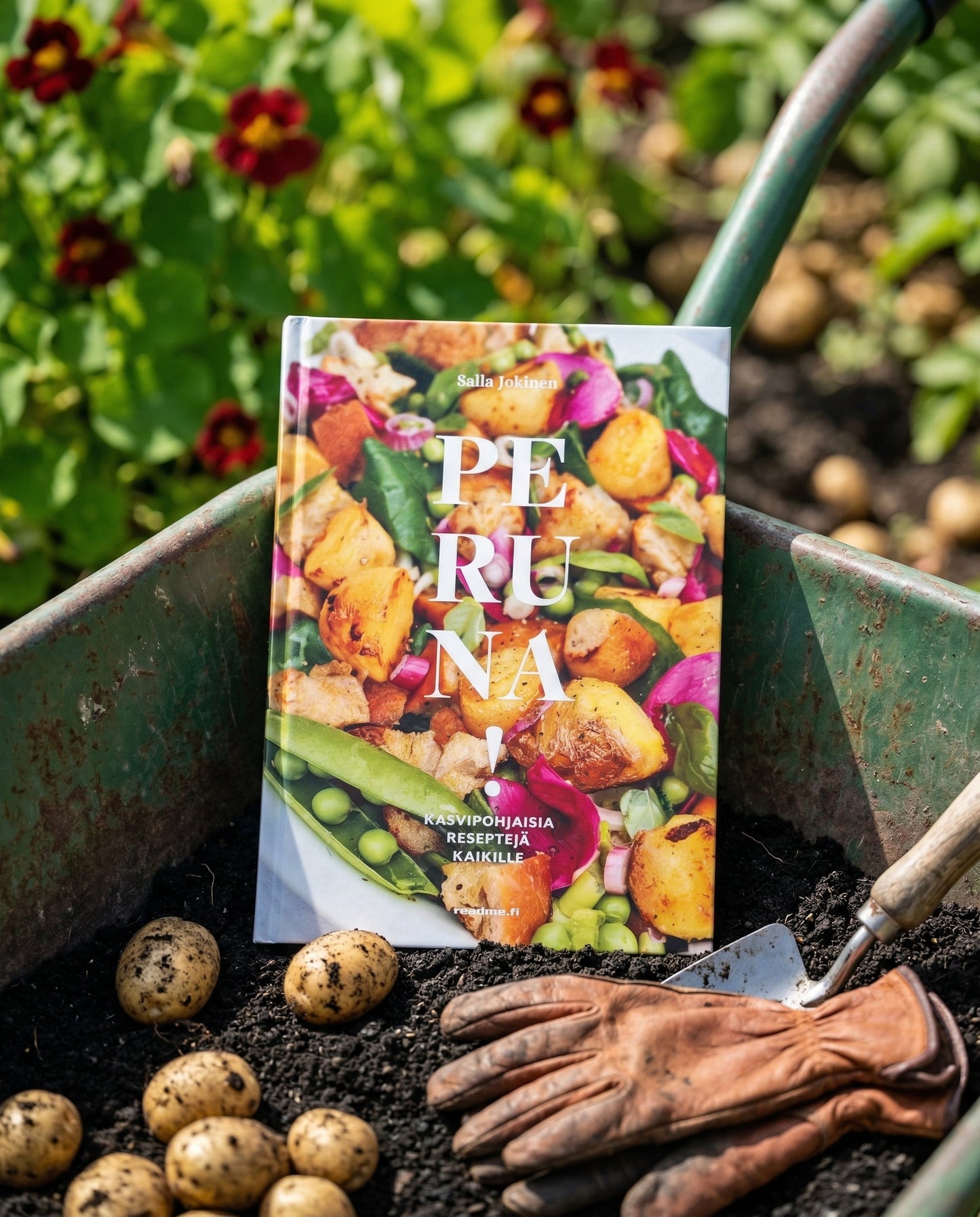 Kottikärryissä on kirja Peruna!, tekijä Salla Jokinen. Kirjan ympärillä on multaa, perunoita, puutarhahanskat ja pieni lapio. Taustalla näkyy vihreitä kasveja ja kukkia.