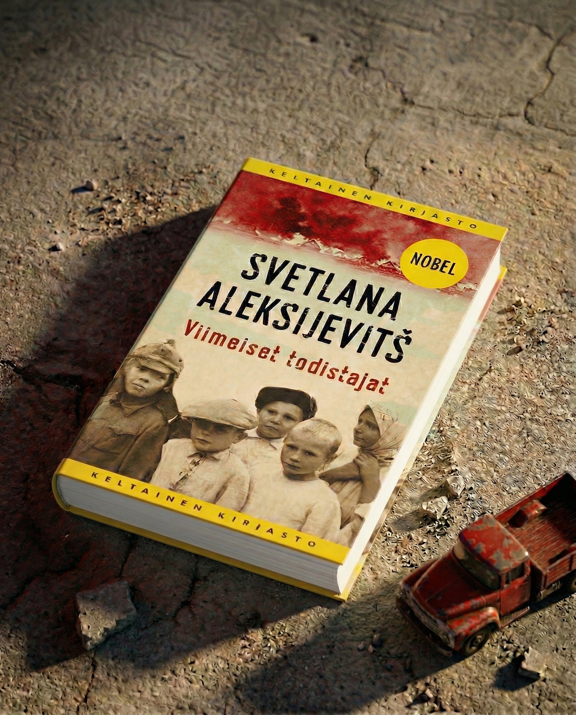 Svetlana Aleksijevitšin kirja Viimeiset todistajat on asetettu karhealle maapinnalle. Kannessa on kuva lapsista ja Nobel-merkintä. Kirjan vieressä on pieni punainen leluauto.